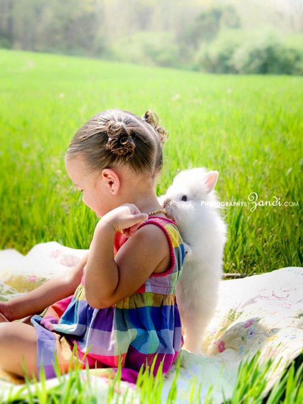 Funny Easter Bunny Picture. The little girl with beautiful dress is sitting on a blanket, the cute bunny is trying to bite her necklace from the back. It’s so wonderful to witness this interesting moment. 